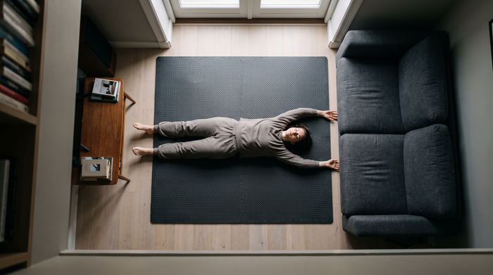 Overhead view of a person lying on a training mat with arms stretched above their head, their body filling the mat's footprint in the narrow gap between a sofa and a desk.